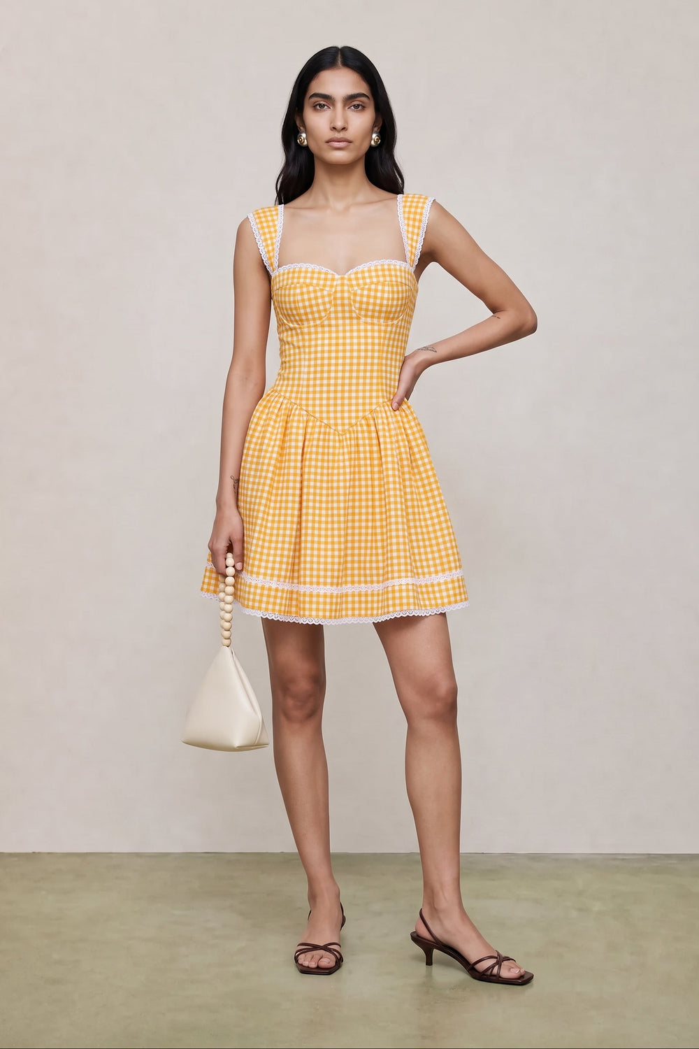 A woman modeling a yellow and white gingham mini dress with a sweetheart neckline, structured bodice, and flared skirt, holding a small cream handbag against a neutral studio backdrop.