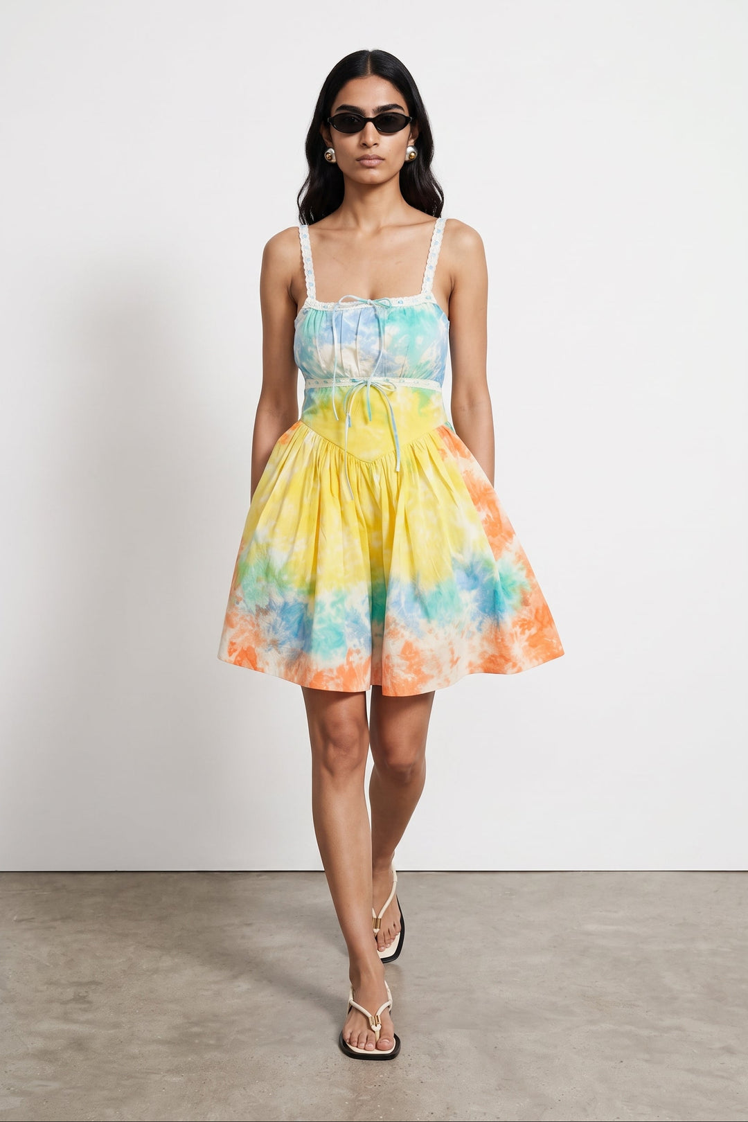 A woman modeling a sleeveless A-line mini dress with a tie-dye pattern in yellow, blue, green, and orange, paired with white spaghetti straps and a matching waist tie, set against a plain white studio wall.