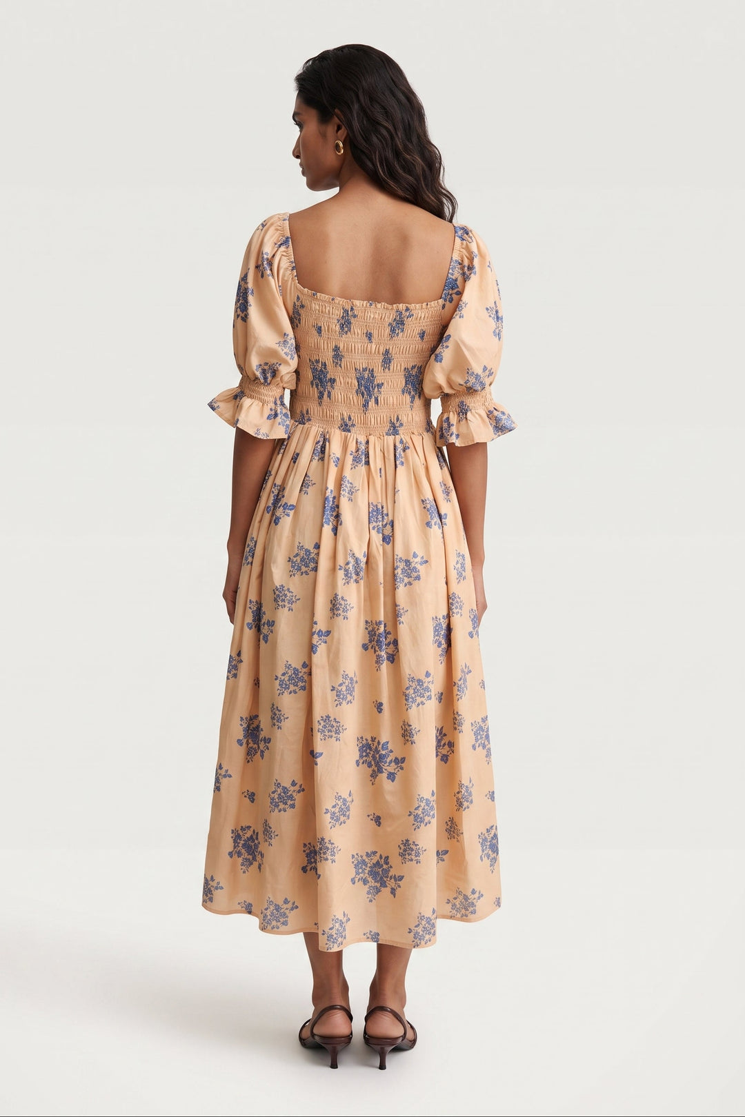 A woman viewed from the back wearing a peach-colored midi dress with a blue floral pattern, puff sleeves, and a square neckline, paired with black heeled sandals against a light gray studio background.