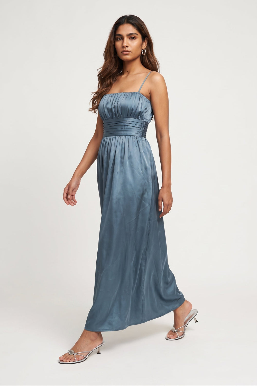 A woman modeling a floor-length, sleeveless blue satin dress with thin straps and a gathered waist, paired with clear high-heeled sandals, against a plain light gray studio background.
