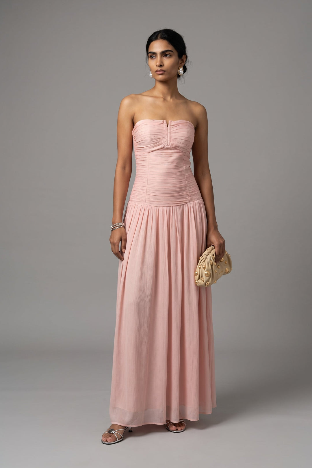 A woman modeling a strapless, floor-length blush pink gown with a ruched bodice and flowing skirt, holding a small woven clutch against a plain gray studio background.