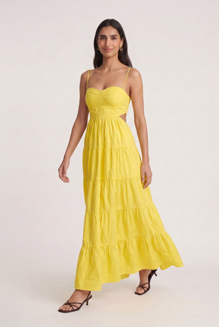 A woman modeling a bright yellow tiered maxi dress with spaghetti straps and side cutouts, paired with black strappy heels, against a plain light beige studio background.