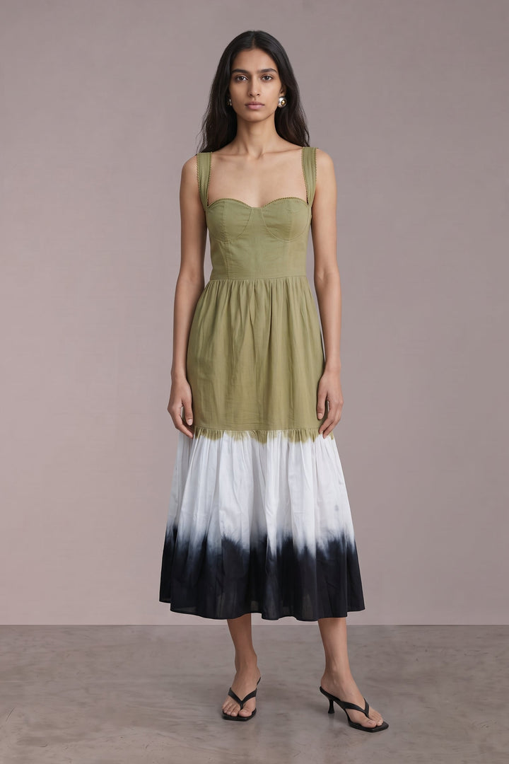 A woman modeling a sleeveless midi dress with an olive green bodice and a white-to-black ombre tie-dye skirt, standing against a solid taupe studio backdrop.