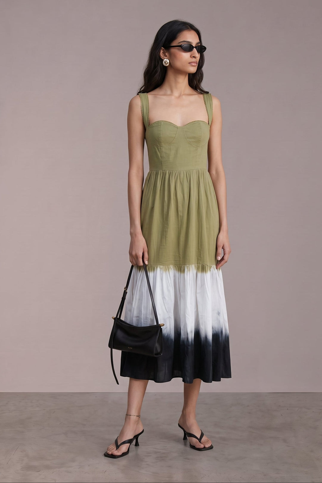 A woman models a sleeveless midi dress with a solid olive green bodice and a white-to-black tie-dye hem, styled with a black handbag and sunglasses against a neutral studio backdrop.