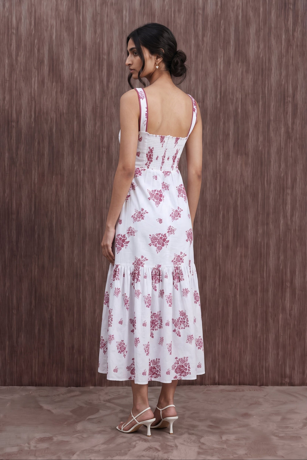 A woman wearing a white sleeveless midi dress with a pink floral print, shown from the back against a brown textured wooden wall.