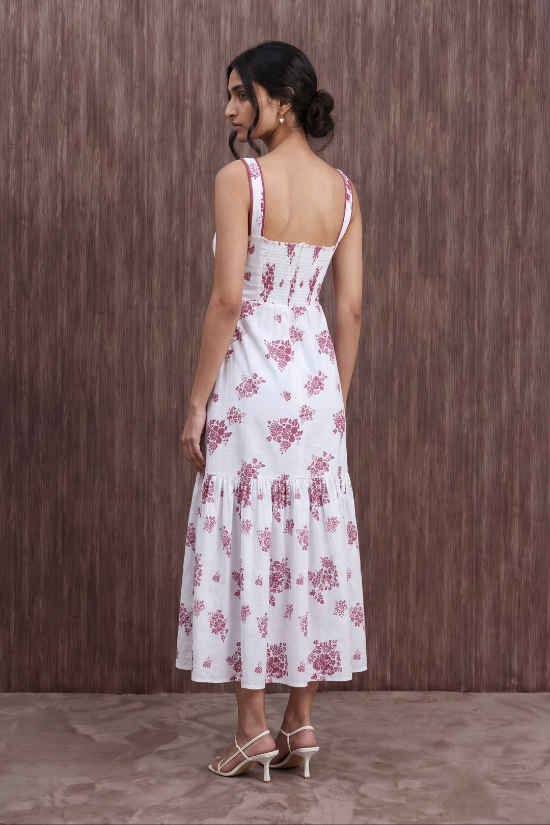 A woman wearing a white sleeveless midi dress with a pink floral print, shown from the back against a brown textured wooden wall.