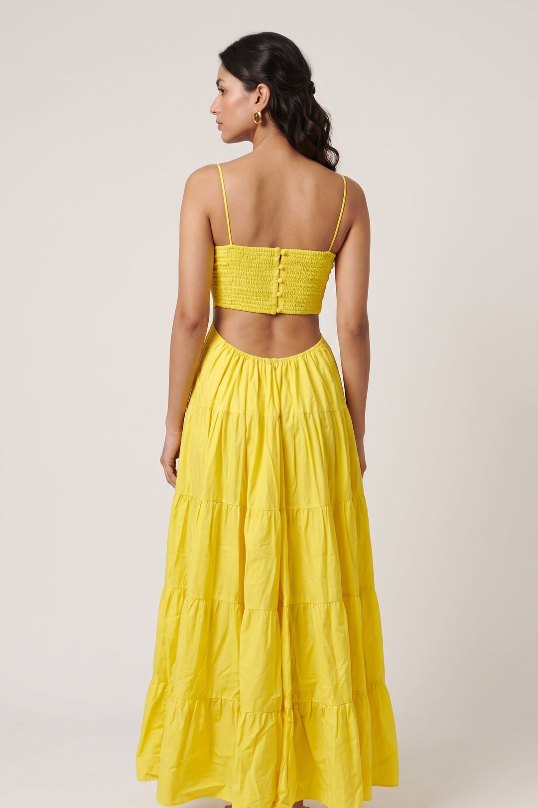A woman shown from the back wearing a bright yellow two-piece outfit consisting of a smocked crop top with thin spaghetti straps and a matching tiered maxi skirt, set against a plain light beige studio background.