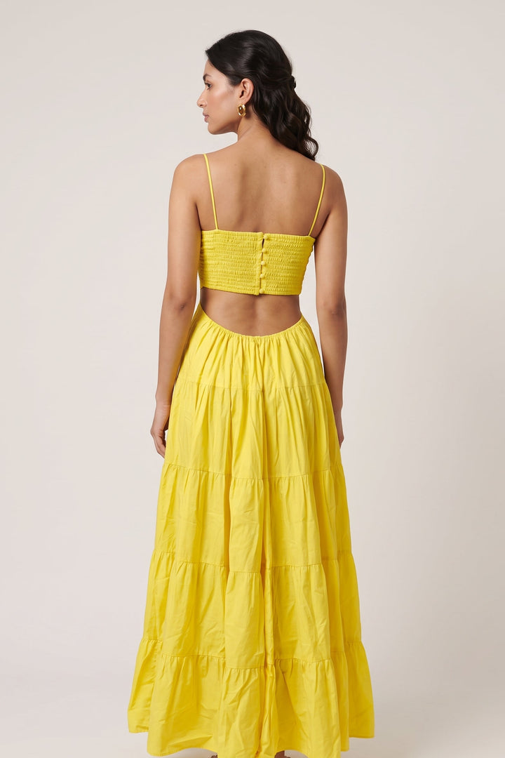 A woman shown from the back wearing a bright yellow two-piece outfit consisting of a smocked crop top with thin spaghetti straps and a matching tiered maxi skirt, set against a plain light beige studio background.