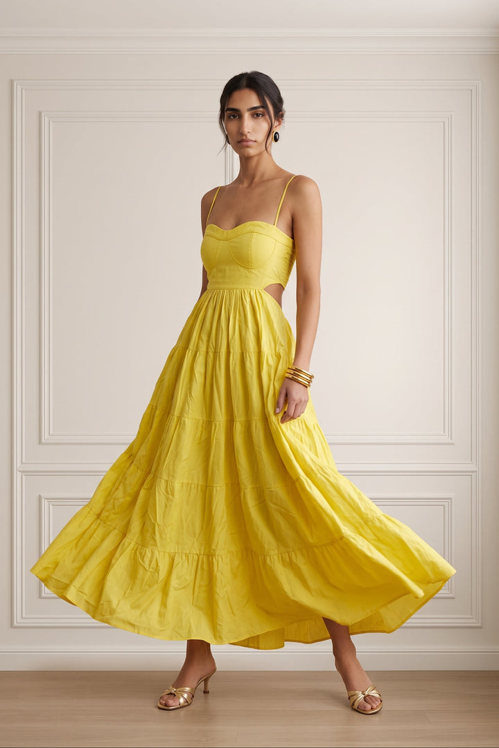 A woman modeling a bright yellow sleeveless maxi dress with thin straps, a fitted bodice, side cutouts, and a full tiered skirt, standing in a cream-colored room with decorative wall paneling.