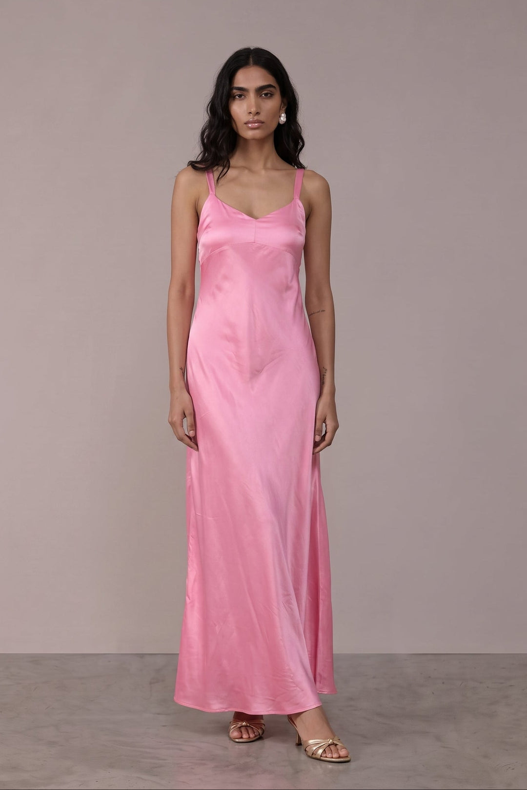 A woman modeling a long, pink satin slip dress with thin spaghetti straps and a V-neckline, standing against a neutral, light gray studio backdrop.