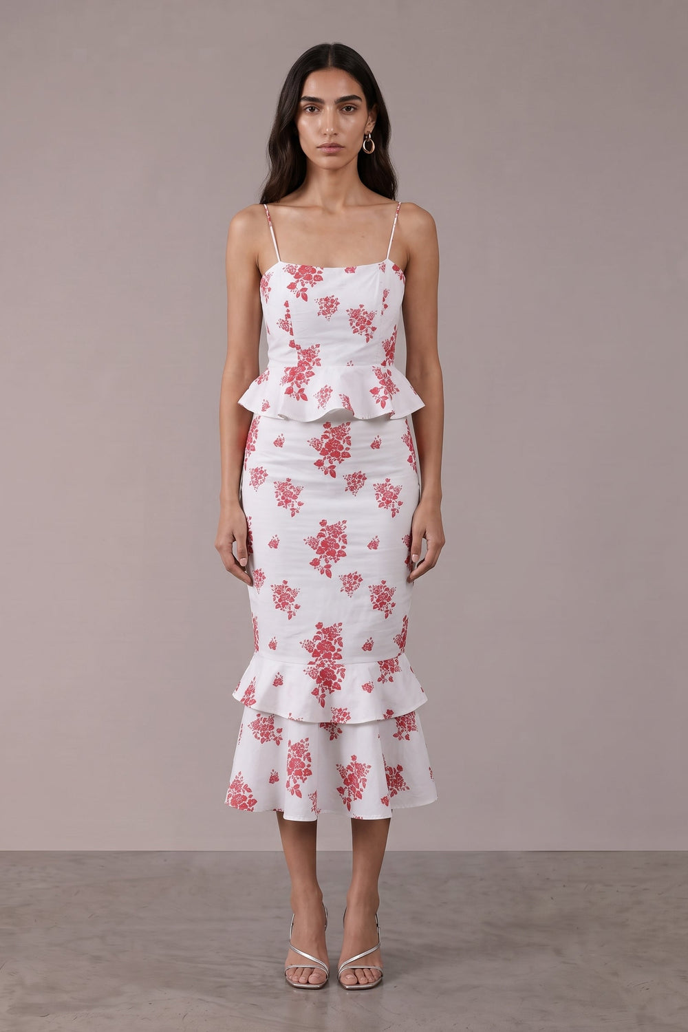 A woman modeling a white midi dress with a pink floral pattern, spaghetti straps, a peplum waist, and a ruffled hem, set against a neutral studio backdrop.
