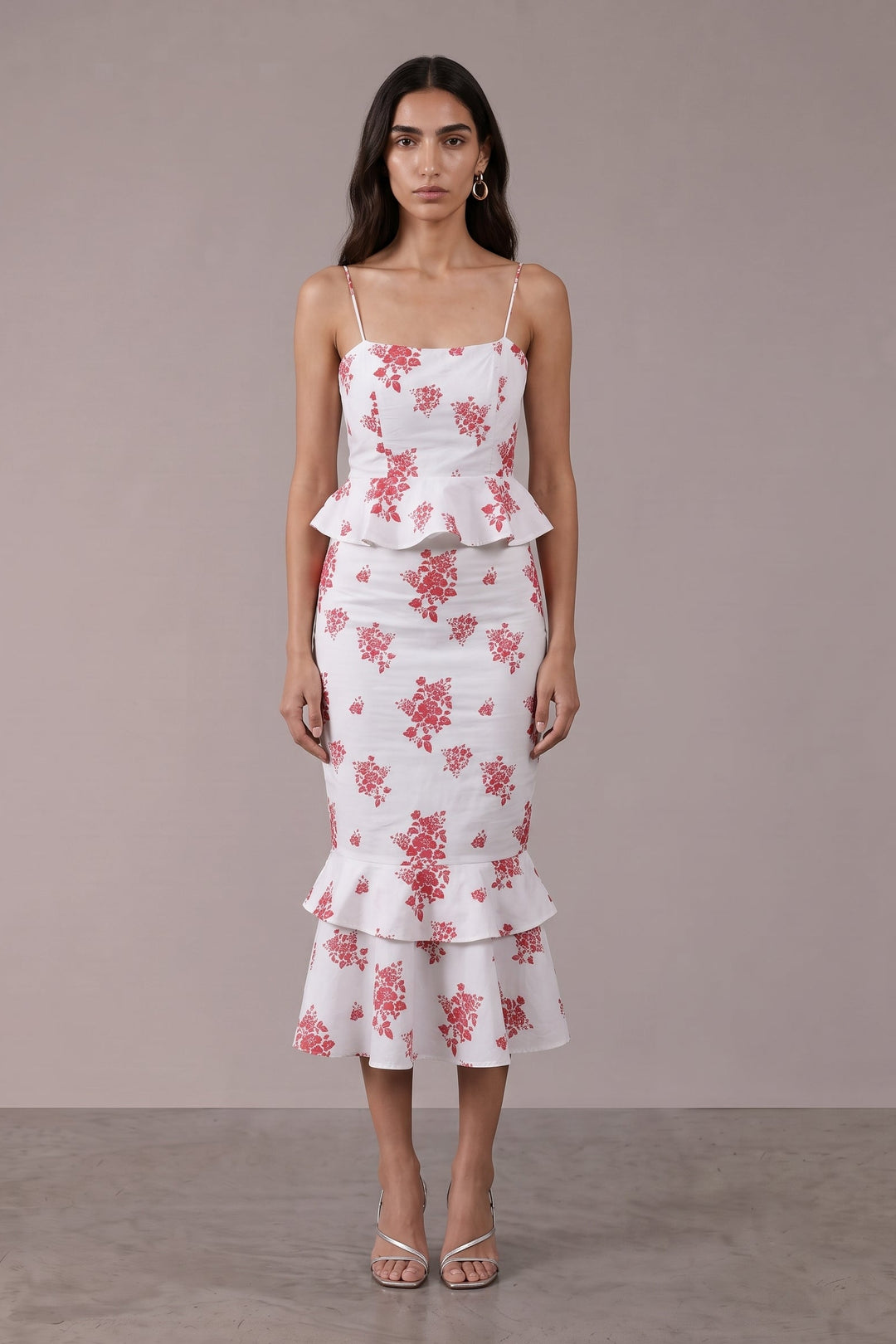 A woman modeling a white midi dress with a pink floral pattern, spaghetti straps, a peplum waist, and a ruffled hem, set against a neutral studio backdrop.