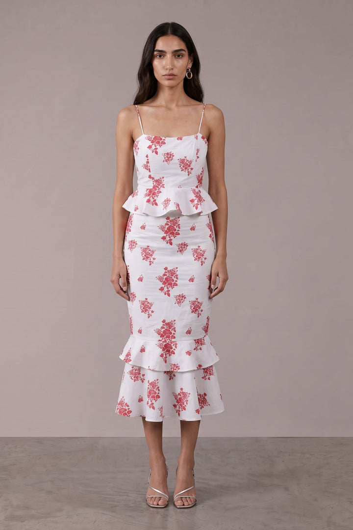 A woman modeling a white midi dress with a pink floral pattern, spaghetti straps, a peplum waist, and a ruffled hem, set against a neutral studio backdrop.