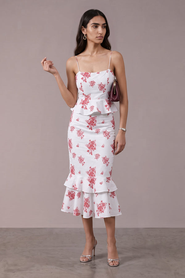 A woman models a white two-piece set featuring a floral print in pink and red tones. The set includes a sleeveless peplum-style top with thin spaghetti straps and a matching tiered ruffle midi skirt.