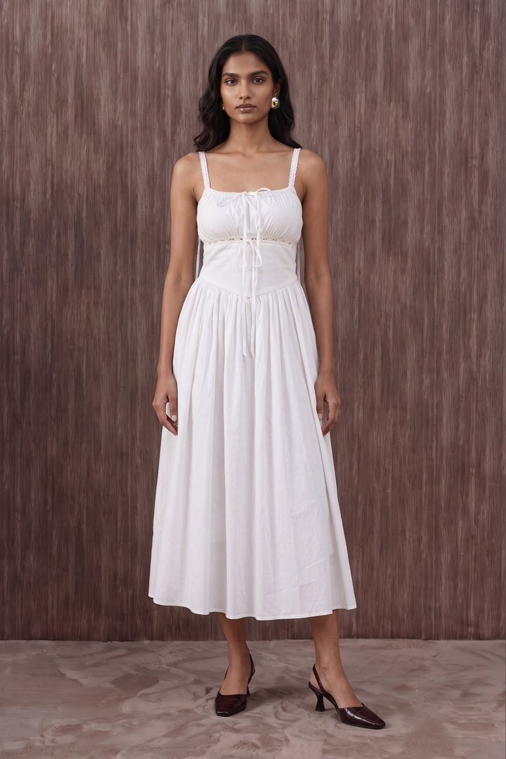 A woman modeling a white midi-length spaghetti-strap dress with a tie-front bodice and gathered skirt, standing against a brown textured wall.