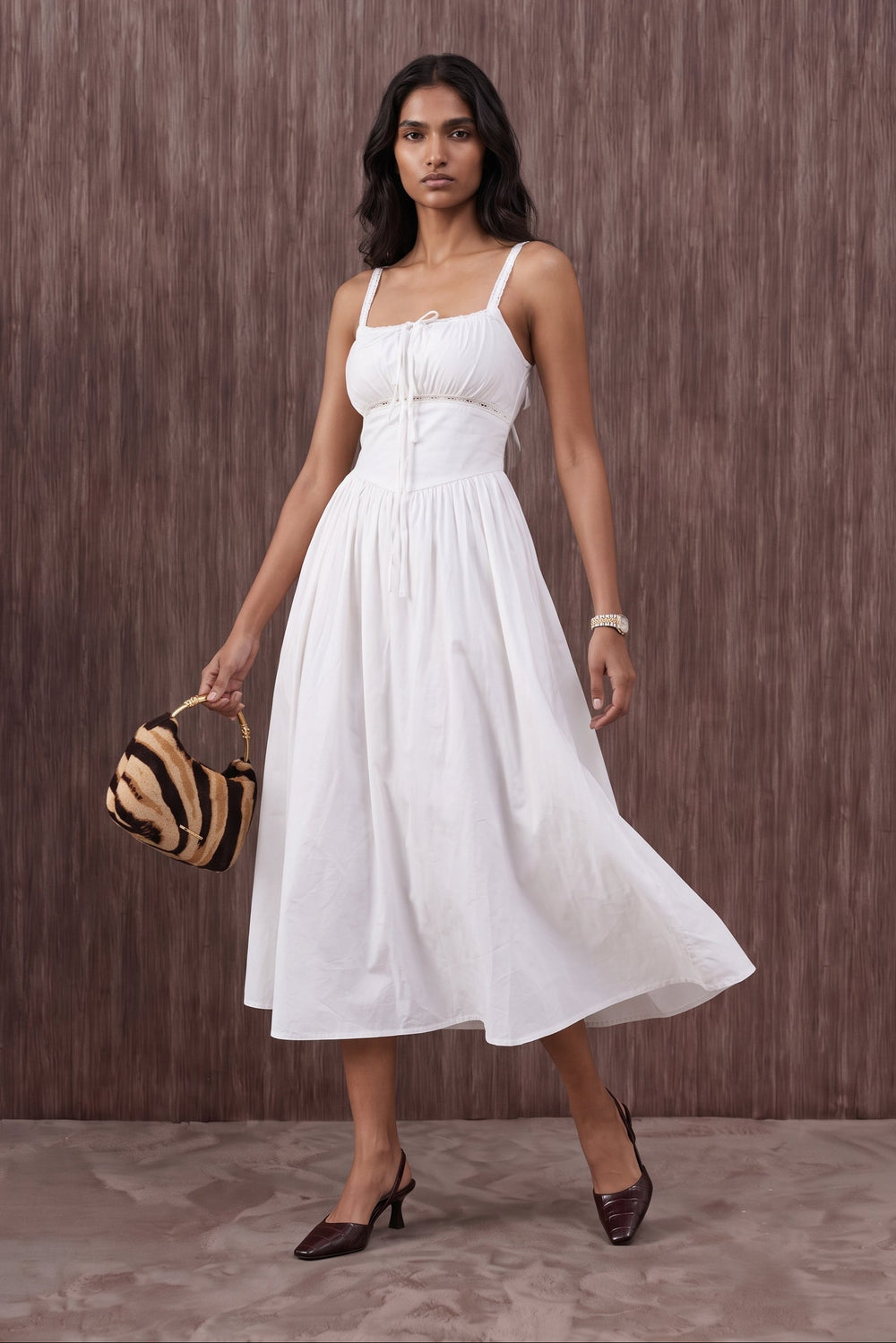 A woman modeling a white sleeveless midi dress with thin straps, a gathered waist, and a flowing skirt, standing against a brown textured wall and holding a small striped handbag.
