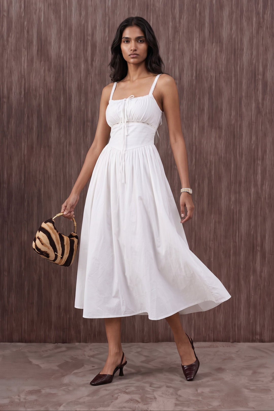 A woman modeling a white sleeveless midi dress with thin straps, a gathered waist, and a flowing skirt, standing against a brown textured wall and holding a small striped handbag.