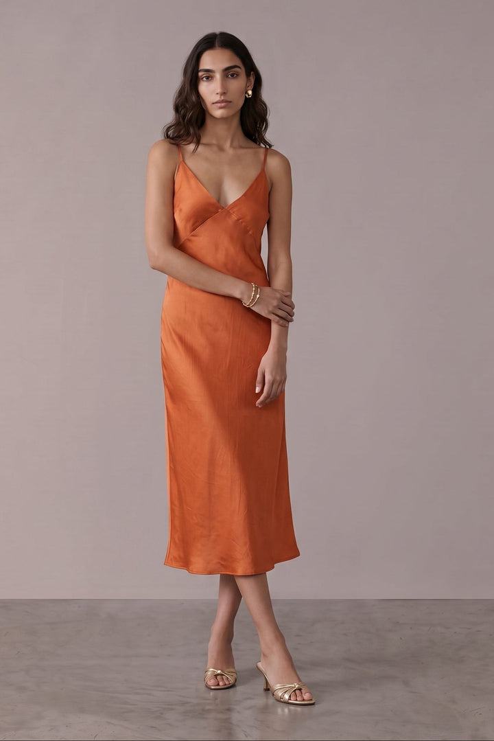 A woman modeling a sleeveless orange midi slip dress with thin straps and a wrap-style bodice, standing against a plain taupe studio backdrop.