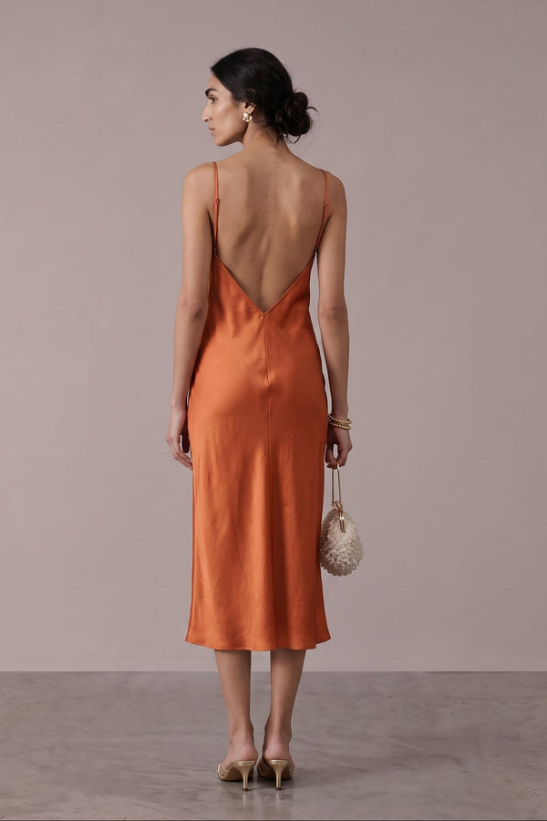 A woman stands with her back to the camera wearing a sleeveless, open-back midi dress in a warm rust-orange color. She is holding a small woven handbag and wearing beige heels, set against a minimalist studio backdrop with a dusty pink wall and gray floor.