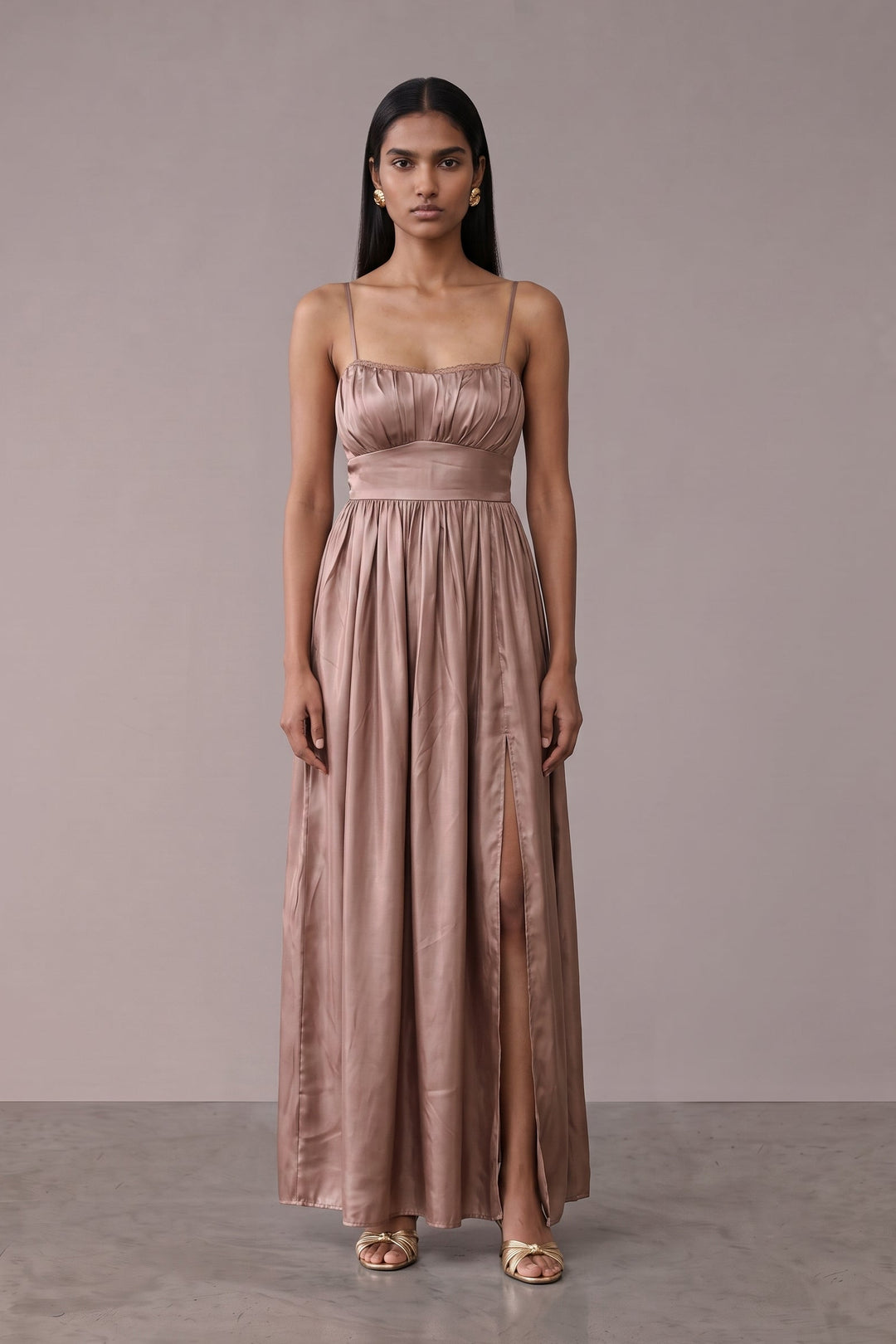 A woman modeling a floor-length dusty rose satin gown with spaghetti straps and a gathered waist, standing against a neutral taupe studio backdrop.