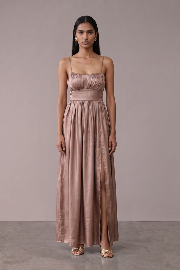 A woman modeling a floor-length dusty rose satin gown with spaghetti straps and a gathered waist, standing against a neutral taupe studio backdrop.