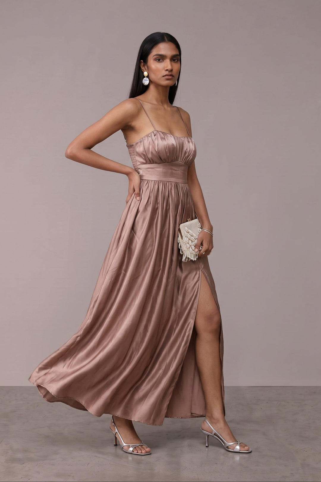 A woman models a floor-length satin strapless gown in a dusty rose pink color, featuring a ruched bodice, thin shoulder straps, a high thigh slit, and a flowing skirt. She is holding a small white beaded clutch and wearing silver high-heeled sandals against a neutral studio backdrop.