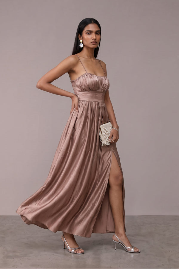 A woman models a floor-length satin strapless gown in a dusty rose pink color, featuring a ruched bodice, thin shoulder straps, a high thigh slit, and a flowing skirt. She is holding a small white beaded clutch and wearing silver high-heeled sandals against a neutral studio backdrop.