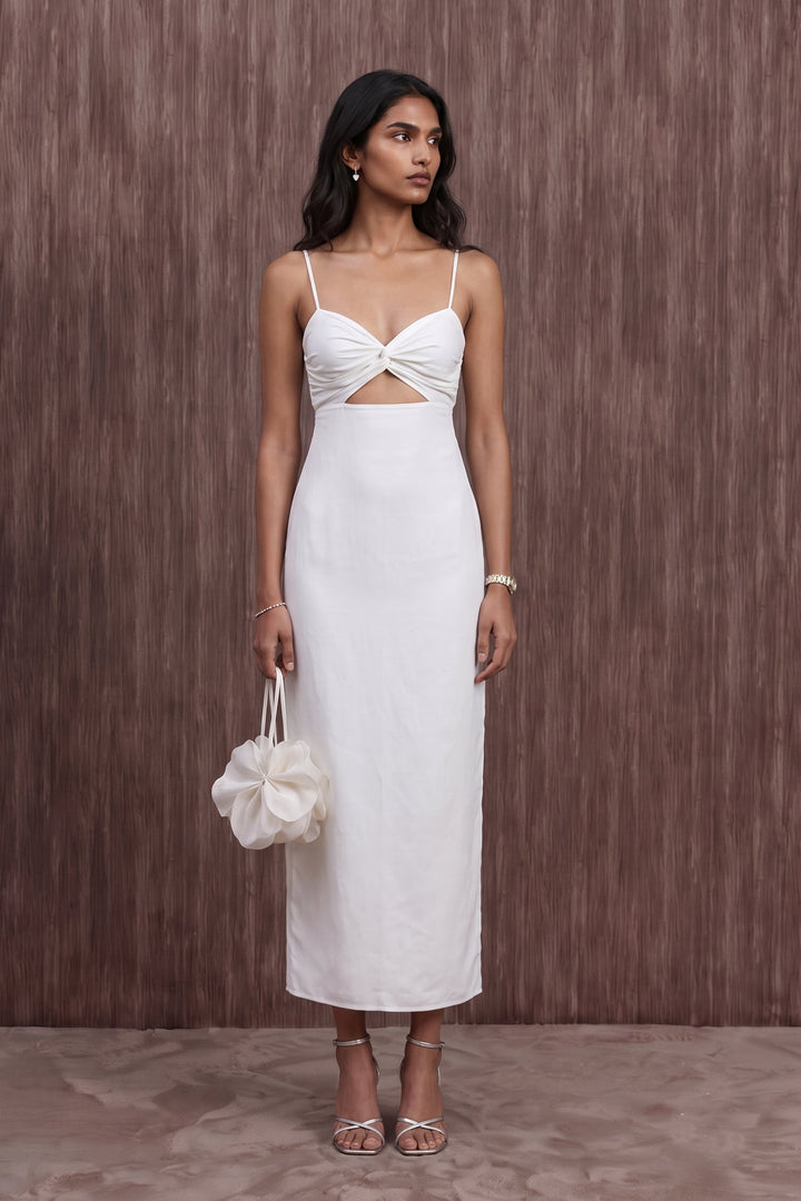 A woman modeling a white midi dress with thin straps, a twisted cutout at the bust, and a fitted skirt, standing against a brown textured wall and holding a matching white handbag.