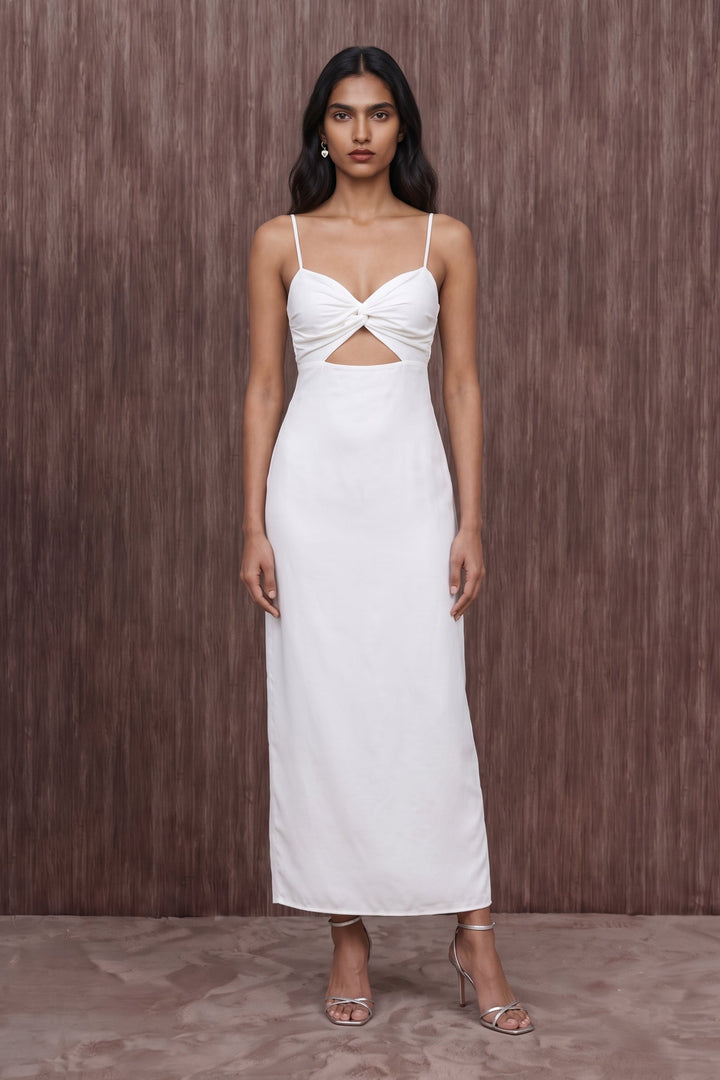 A woman modeling a white sleeveless maxi dress with spaghetti straps, a twisted sweetheart neckline, and a front cutout, standing against a brown textured wall.