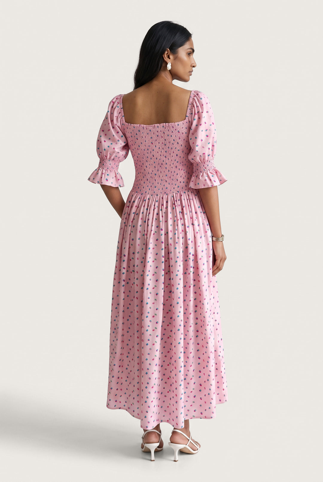 A woman modeling a pink midi dress with a small blue and yellow floral print, featuring puff sleeves and a square neckline, shown from the back against a light beige studio background.