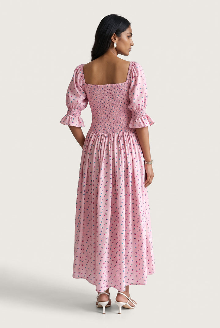 A woman modeling a pink midi dress with a small blue and yellow floral print, featuring puff sleeves and a square neckline, shown from the back against a light beige studio background.