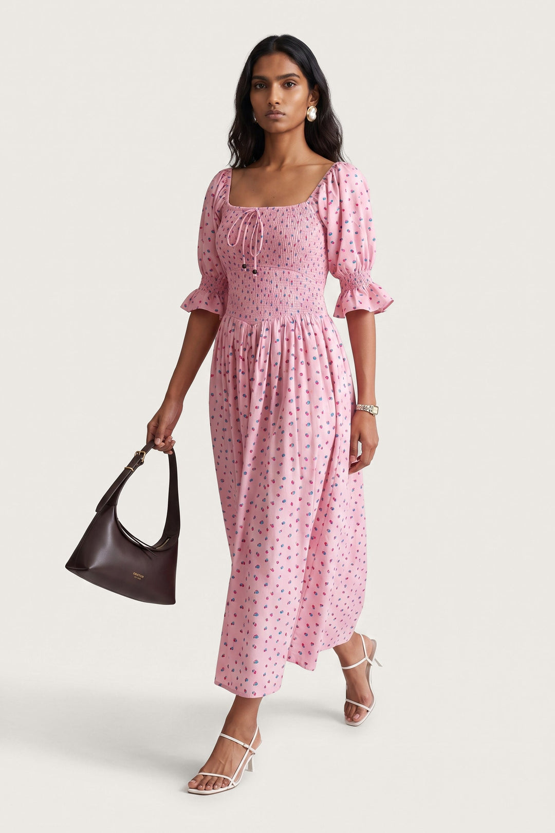 A woman models a pink midi dress with a small red and blue floral print, featuring puff sleeves and a square neckline. She is holding a dark brown leather handbag and wearing white strappy high-heeled sandals against a light beige studio background.