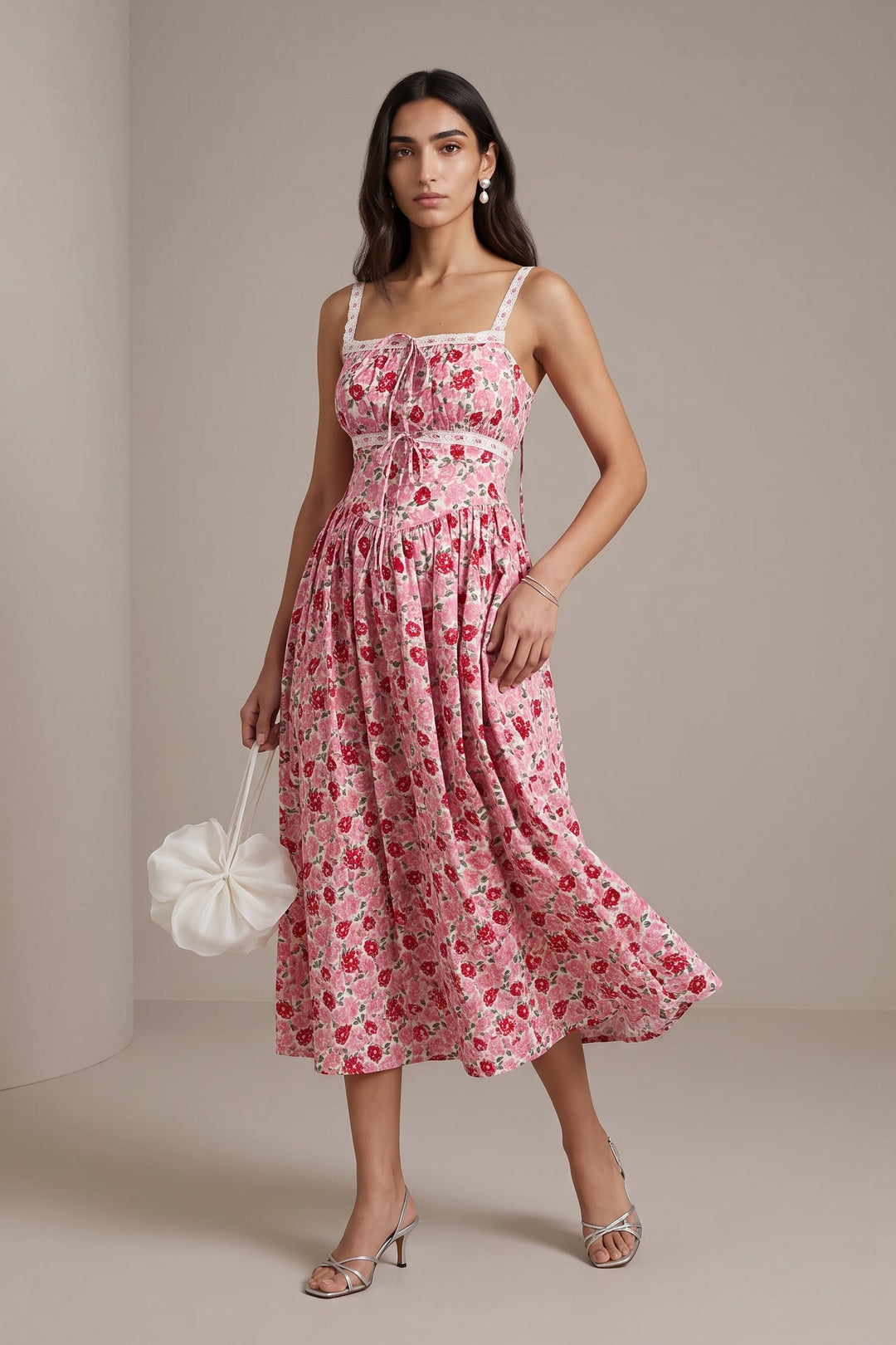 A woman models a pink floral midi dress with white spaghetti straps and a gathered waist, holding a white ruffled handbag against a neutral beige studio backdrop.
