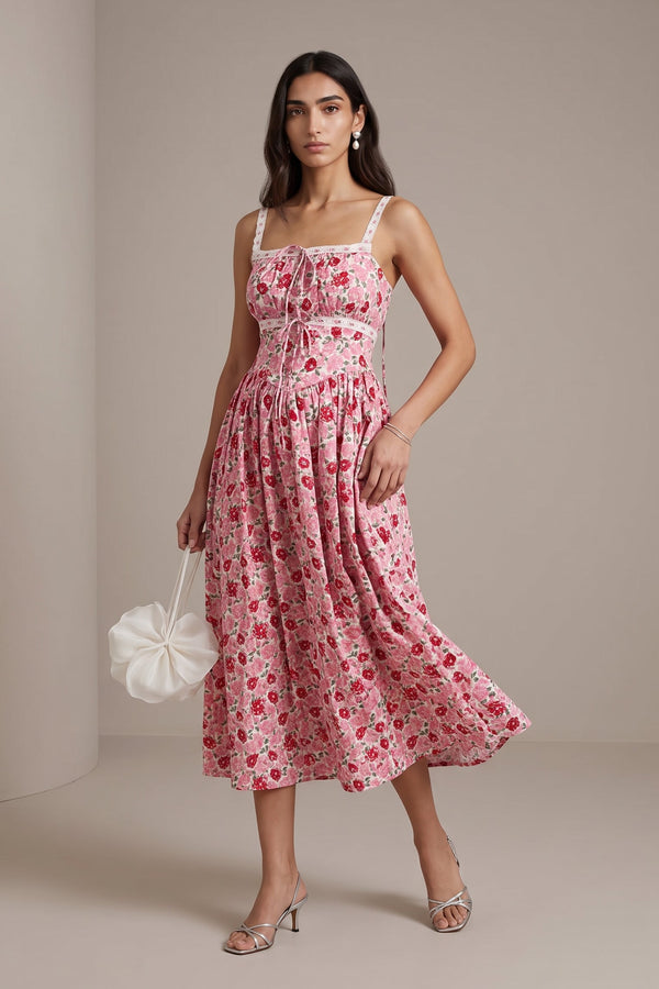 A woman models a pink floral midi dress with white spaghetti straps and a gathered waist, holding a white ruffled handbag against a neutral beige studio backdrop.