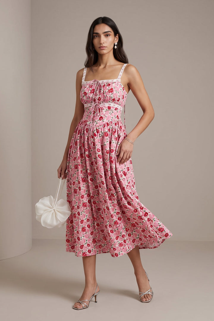 A woman models a pink floral midi dress with white spaghetti straps and a gathered waist, holding a white ruffled handbag against a neutral beige studio backdrop.