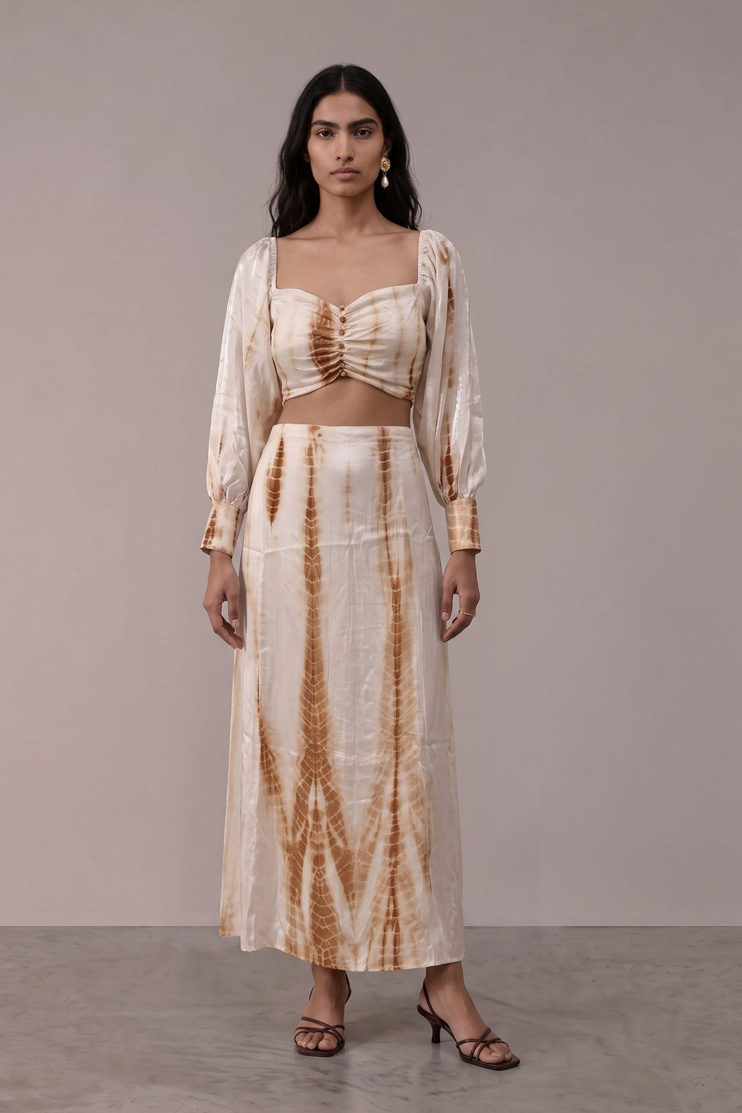 A woman modeling a two-piece tie-dye outfit consisting of a cropped long-sleeve top and a matching maxi skirt, in a light beige color with brown tie-dye patterns, set against a plain studio backdrop.