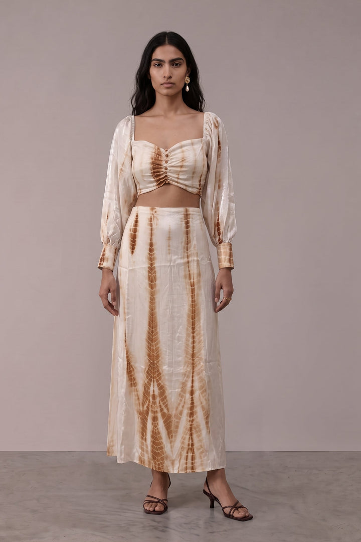 A woman modeling a two-piece tie-dye outfit consisting of a cropped long-sleeve top and a matching maxi skirt, in a light beige color with brown tie-dye patterns, set against a plain studio backdrop.