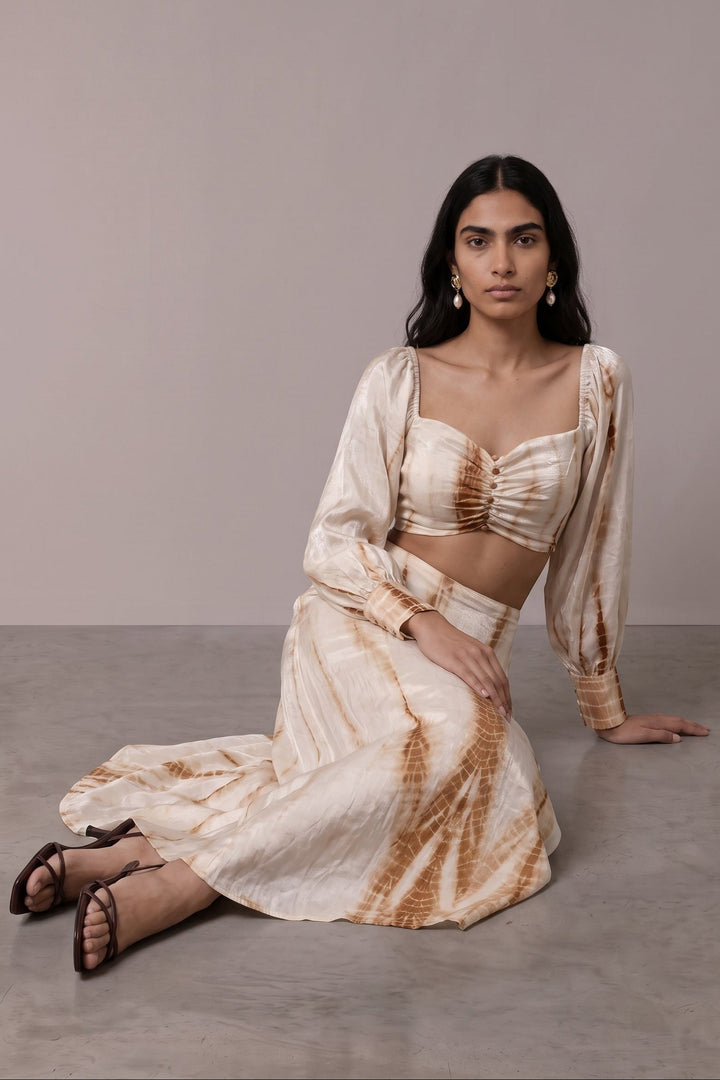 A woman kneels on a gray concrete floor against a neutral beige studio wall, wearing a matching two-piece outfit with a cream and brown tie-dye pattern. The set includes a cropped long-sleeve top with a sweetheart neckline and a long skirt.