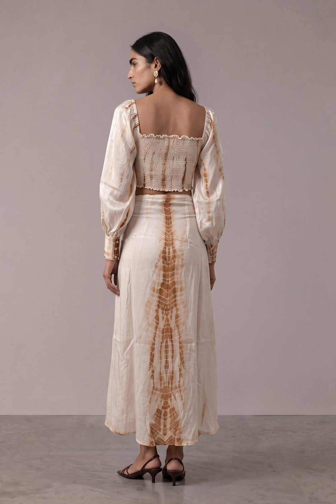 A woman stands with her back to the camera wearing a two-piece cream and brown tie-dye set consisting of a long-sleeve crop top and a matching maxi skirt, set against a plain studio backdrop.