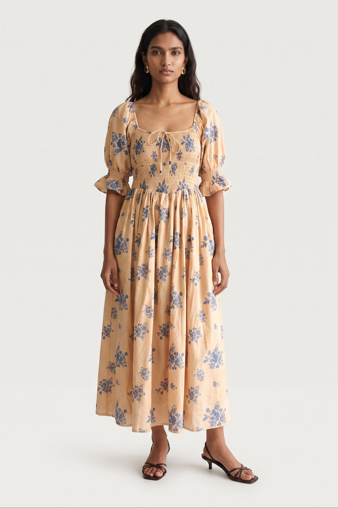 A woman modeling a midi-length cream dress with a blue floral print, featuring puff sleeves, a smocked bodice, and a tie-neck detail, set against a plain light gray background.