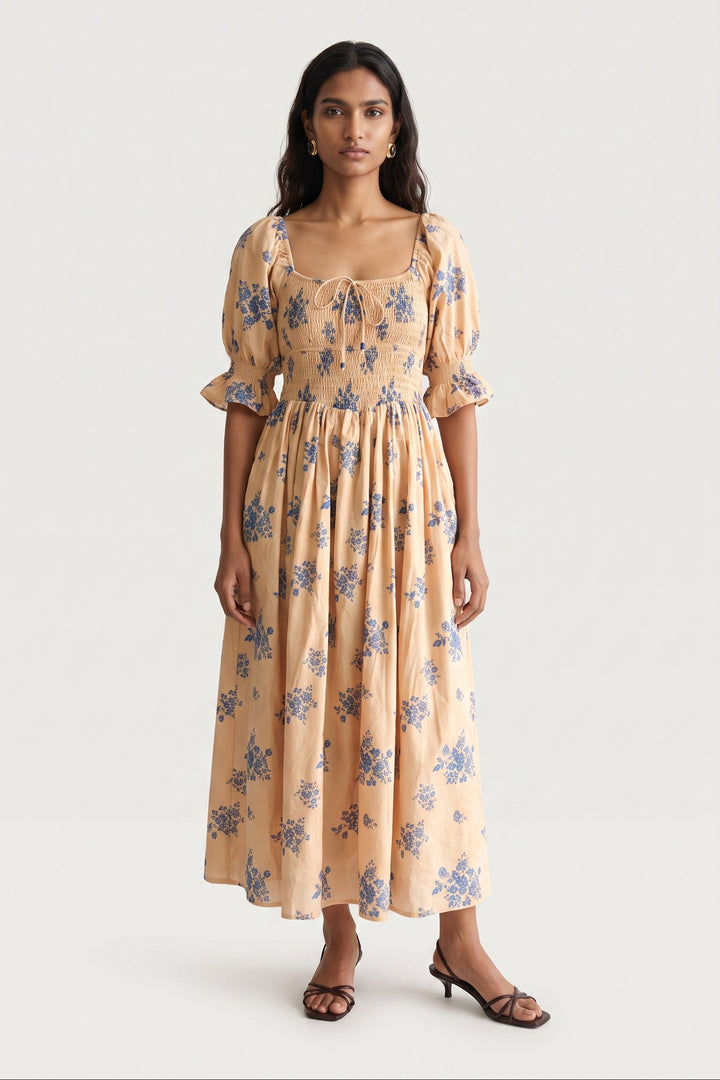 A woman modeling a midi-length cream dress with a blue floral print, featuring puff sleeves, a smocked bodice, and a tie-neck detail, set against a plain light gray background.