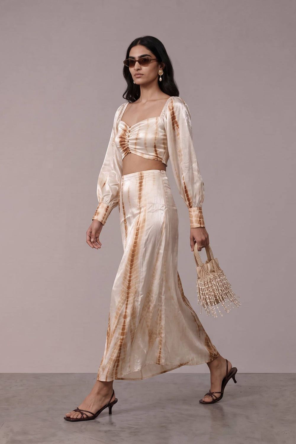 A woman models a two-piece tie-dye set consisting of a cropped long-sleeve top and a matching maxi skirt, styled with a woven straw bag and black strappy heels against a neutral studio backdrop.