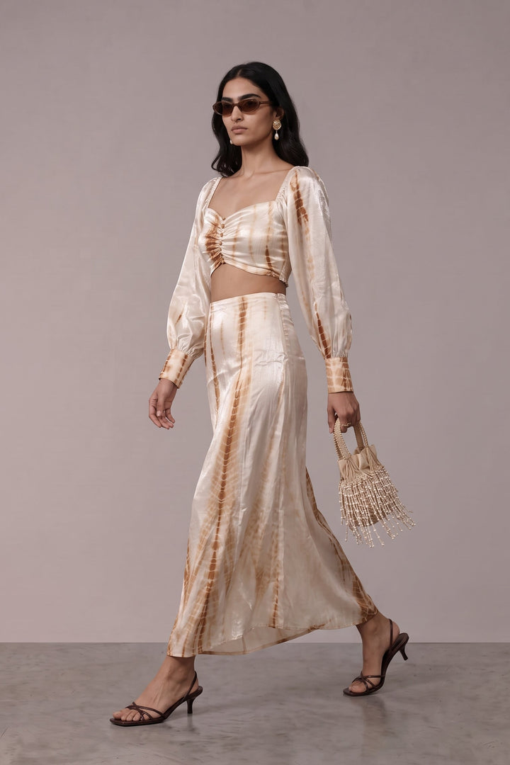 A woman models a two-piece tie-dye set consisting of a cropped long-sleeve top and a matching maxi skirt, styled with a woven straw bag and black strappy heels against a neutral studio backdrop.