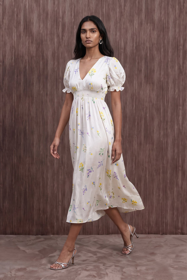 A woman modeling a white midi dress with a yellow and purple floral print, featuring puff sleeves and a V-neckline, set against a brown textured studio backdrop.