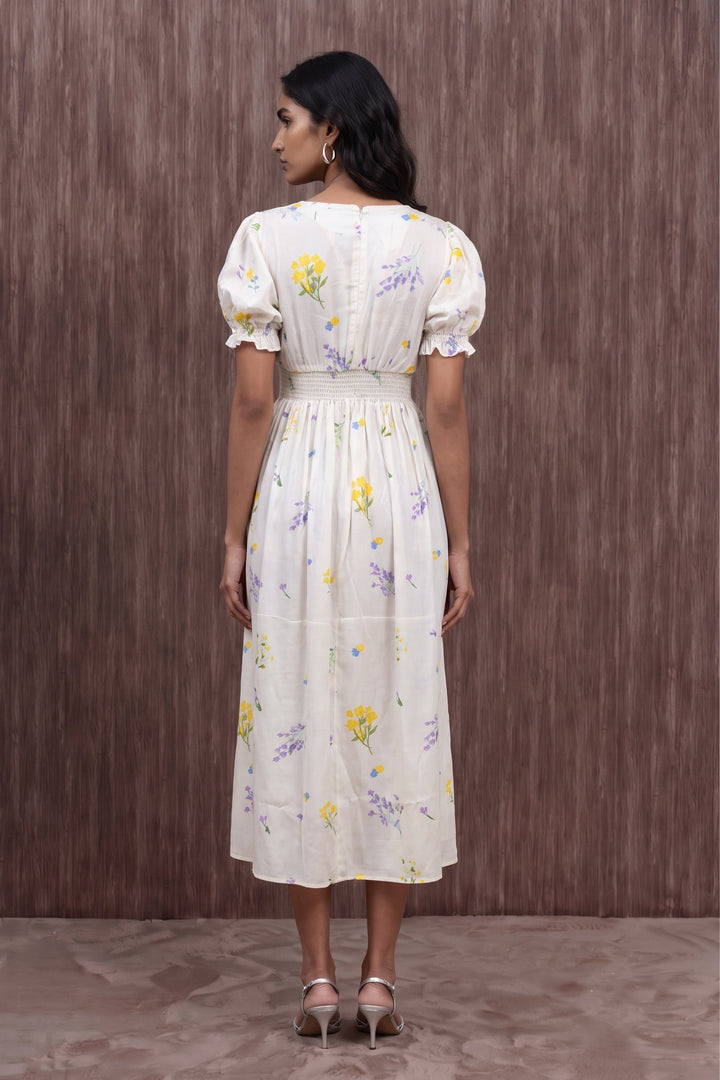 A woman viewed from the back wearing a cream-colored midi dress with puff sleeves, a smocked waist, and a yellow and purple floral print, standing against a brown textured wall.