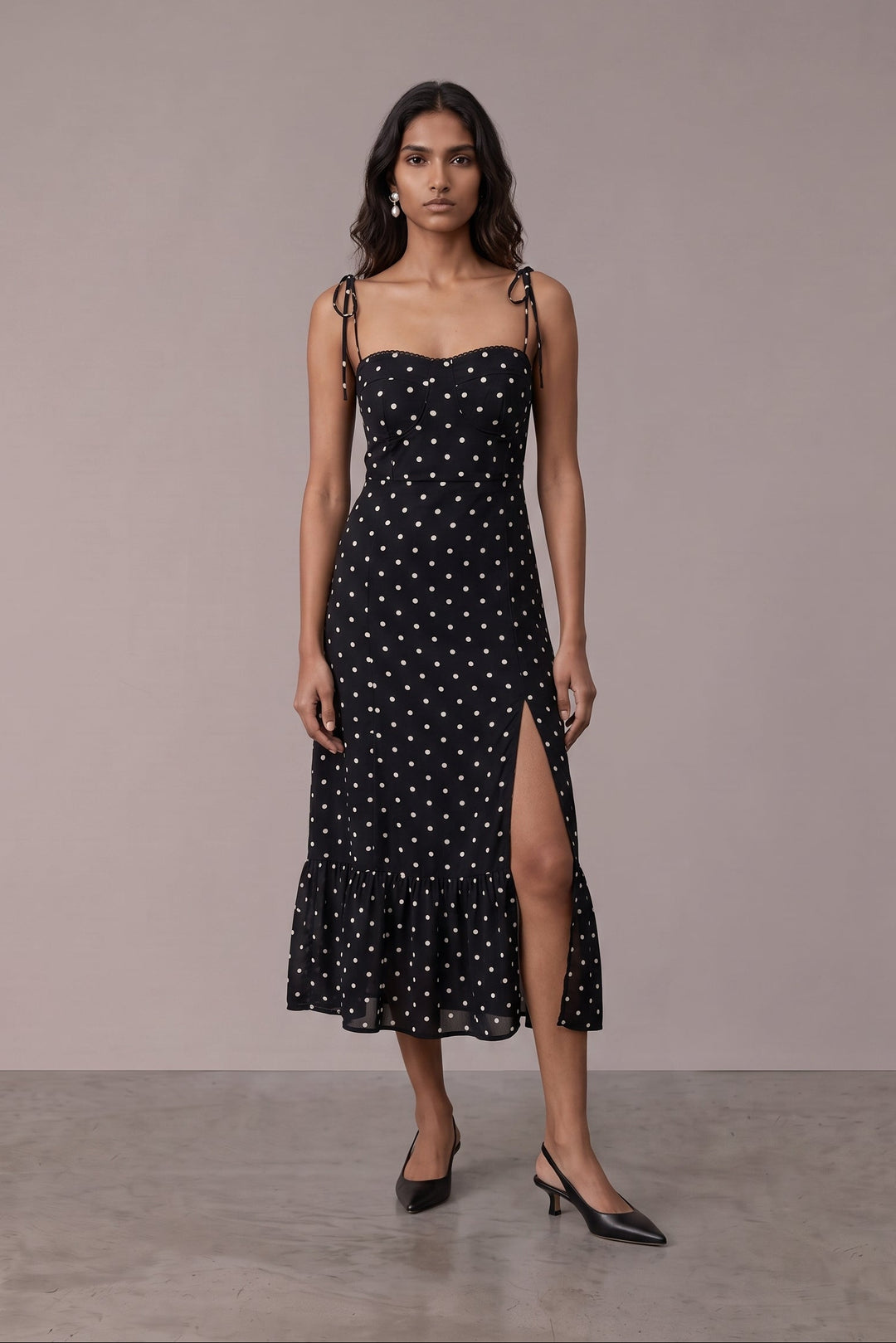 A woman modeling a black midi dress with white polka dots, thin shoulder straps, and a high side slit, standing against a plain beige studio backdrop.