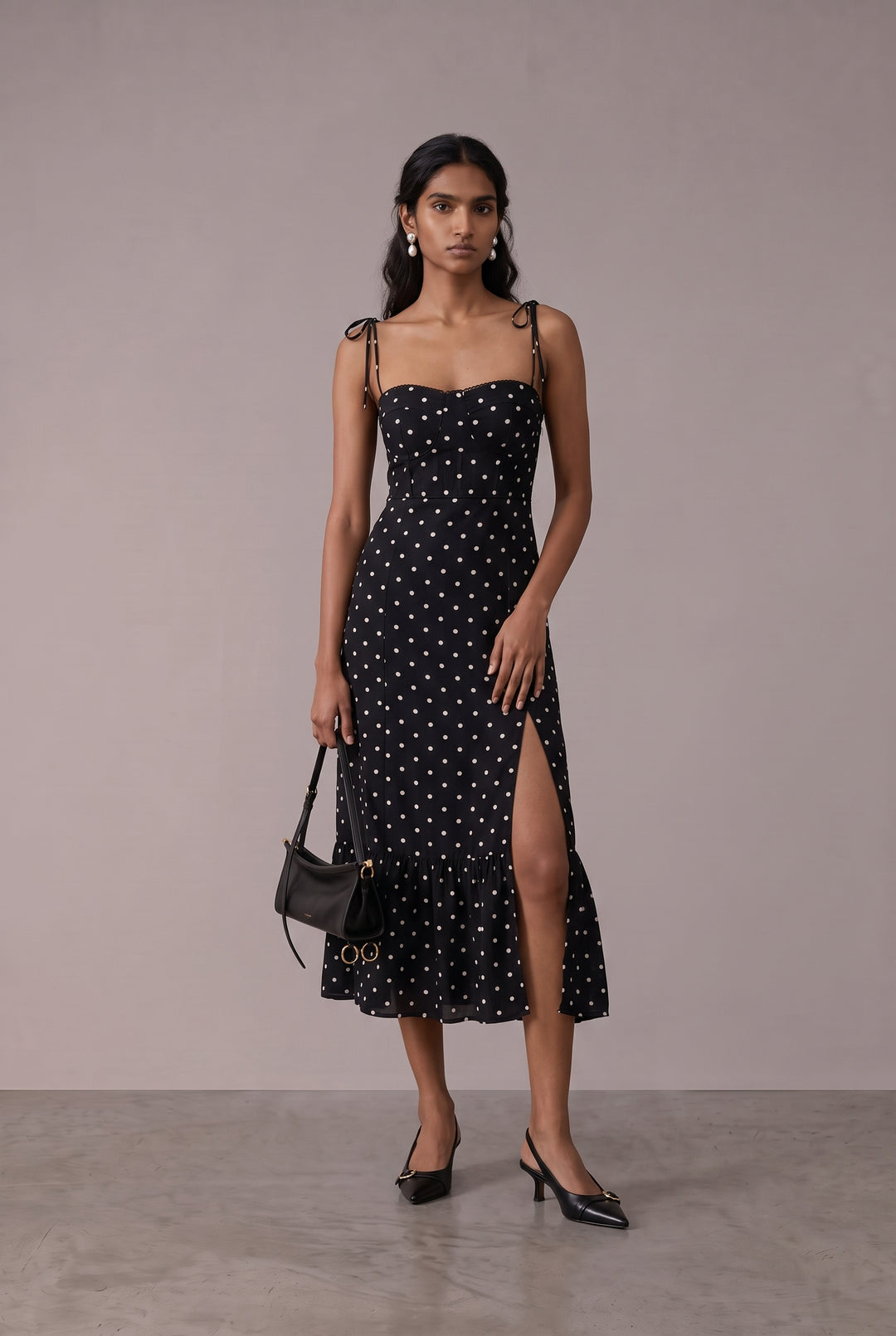 A woman models a black midi dress with white polka dots, featuring thin shoulder straps, a sweetheart neckline, and a high side slit. She is holding a small black handbag and wearing black pointed-toe heels, set against a neutral studio backdrop.