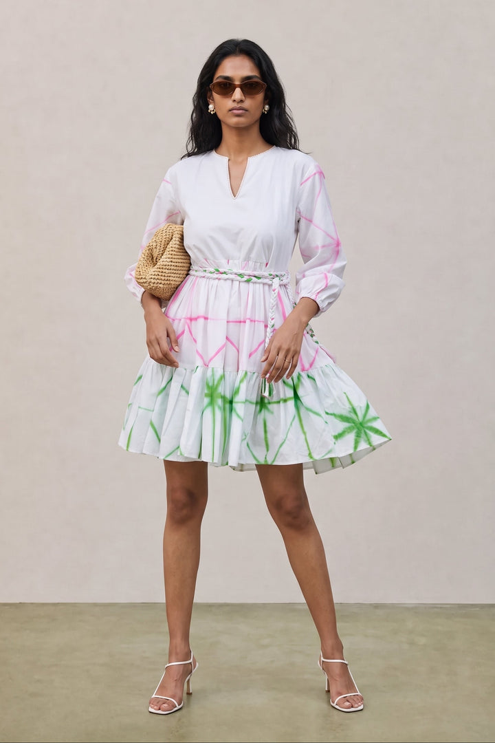 A woman models a white long-sleeve mini dress with pink and green tropical palm tree prints, styled with a woven straw clutch and white strappy heels against a neutral beige background.