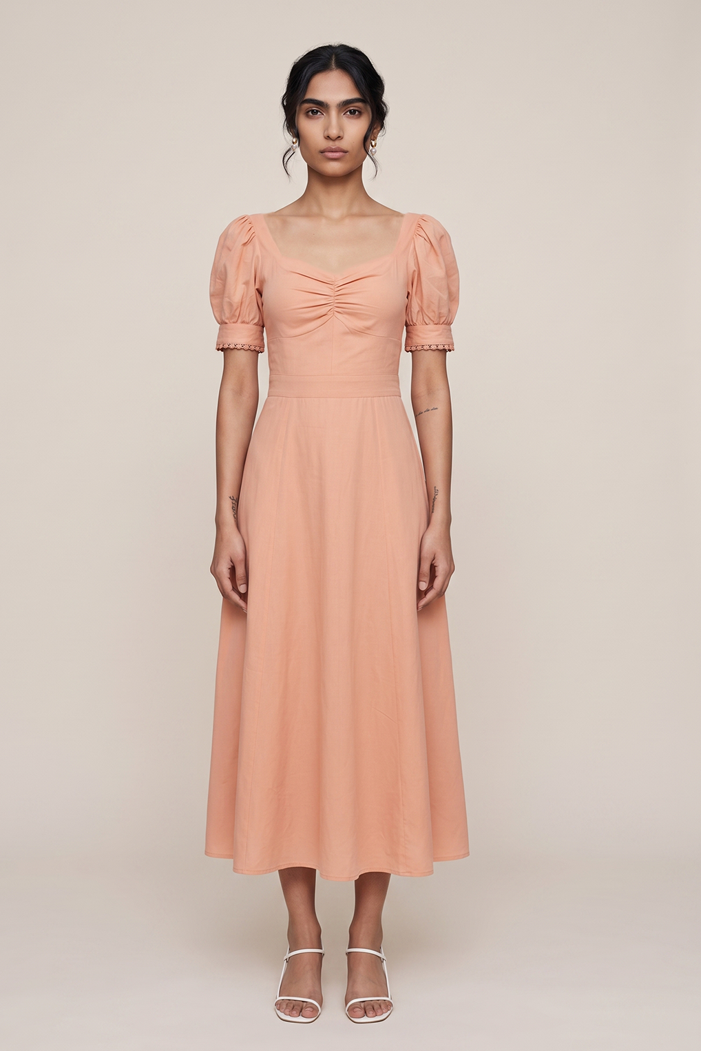 A woman modeling a peach-colored midi dress with puff sleeves, a square neckline, and a gathered front, standing against a plain beige studio background.