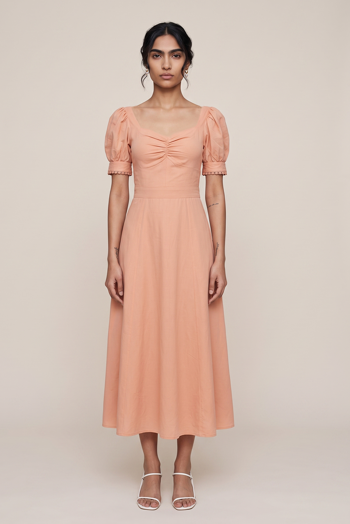 A woman modeling a peach-colored midi dress with puff sleeves, a square neckline, and a gathered front, standing against a plain beige studio background.